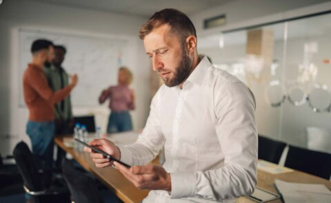 Business man in white shirt no jacket looking at iPad with co-workers behind him blurry