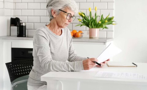 Middle-aged woman in gray sweatshirt and glasses sitting at table looking at papers