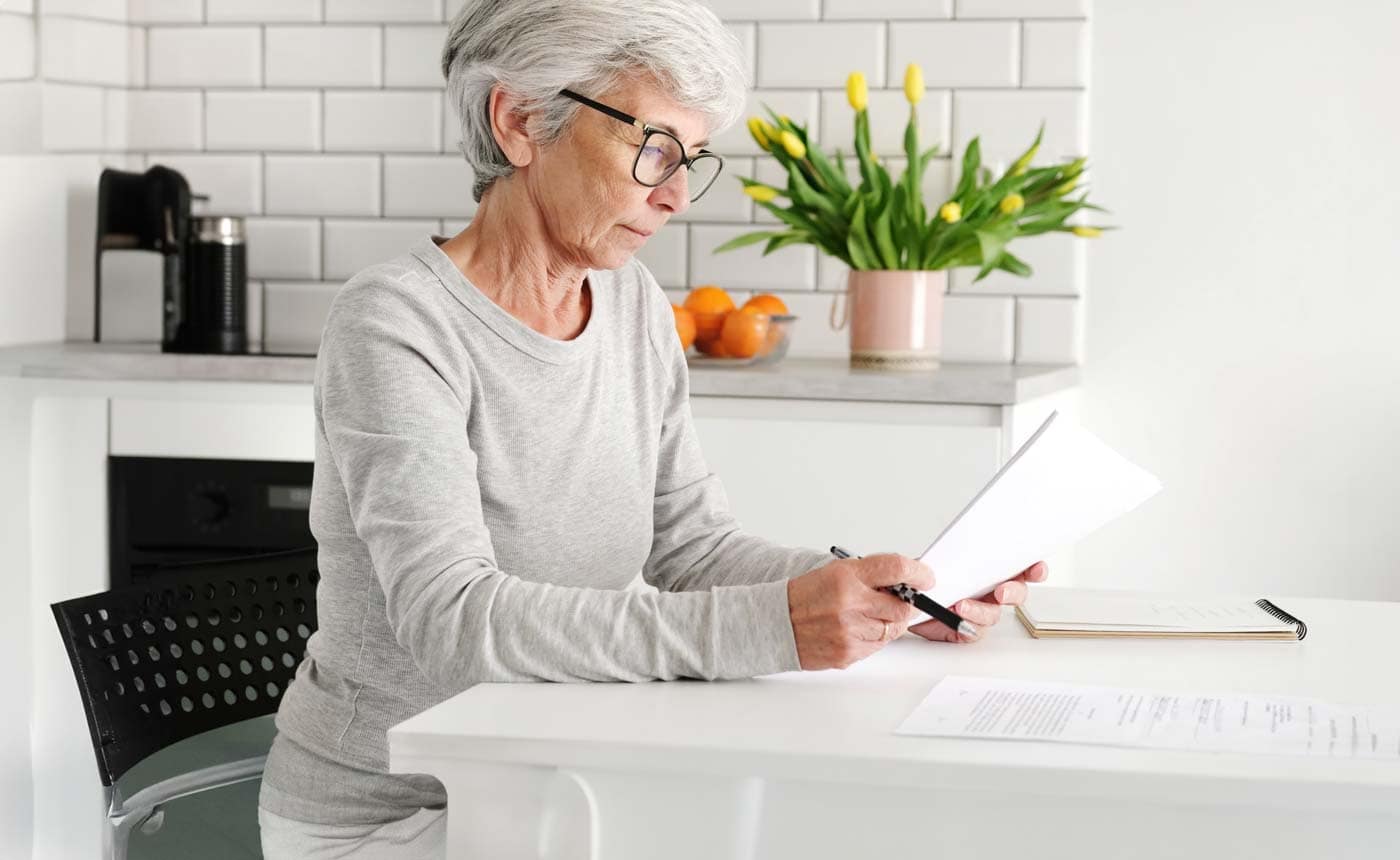 Middle-aged woman in gray sweatshirt and glasses sitting at table looking at papers