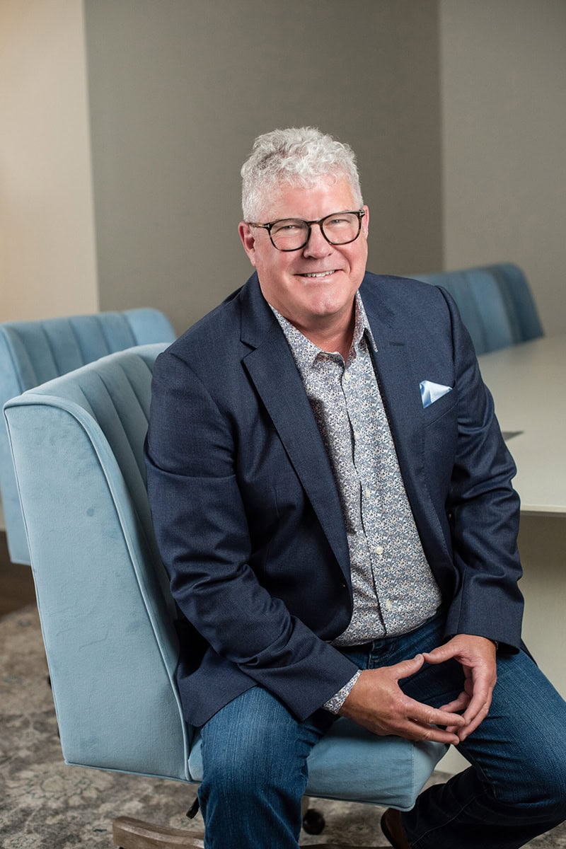 Greg Bennett in a blazer and glasses seated on a blue chair in a professional setting.