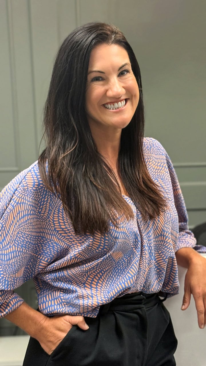 Alexandria Zeis, a woman with long dark hair, wearing a patterned blue and orange blouse and black high-waisted trousers, standing and smiling with one hand in her pocket. A green paneled wall is visible in the background.