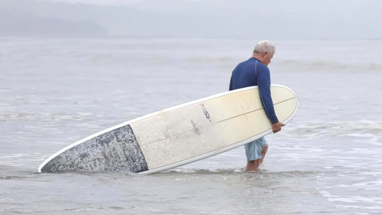 Older man walking into ocean holding surfboard